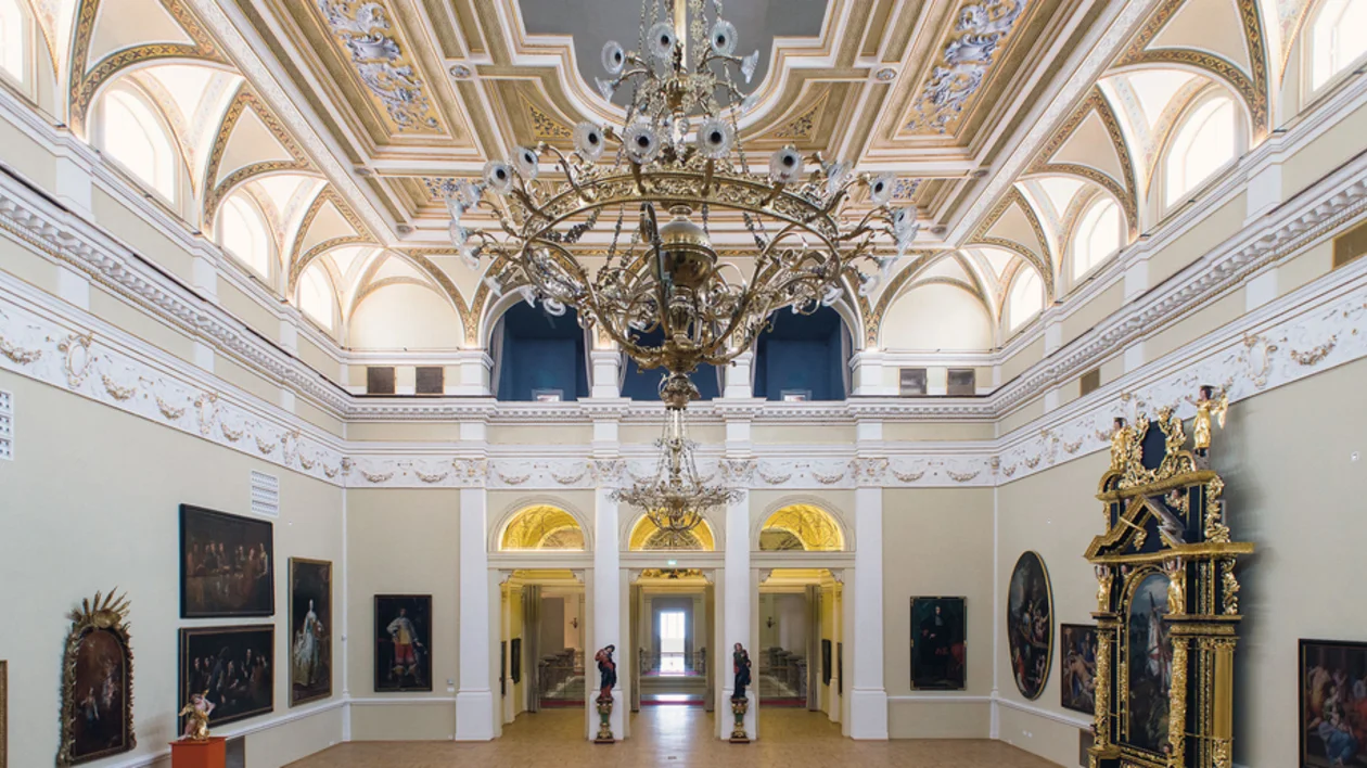 Gallery interior with golden arched ceilings, chandeliers, and classic paintings on walls.