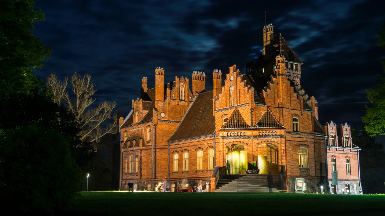 Illuminated brick castle with gothic features at night under a cloudy sky.