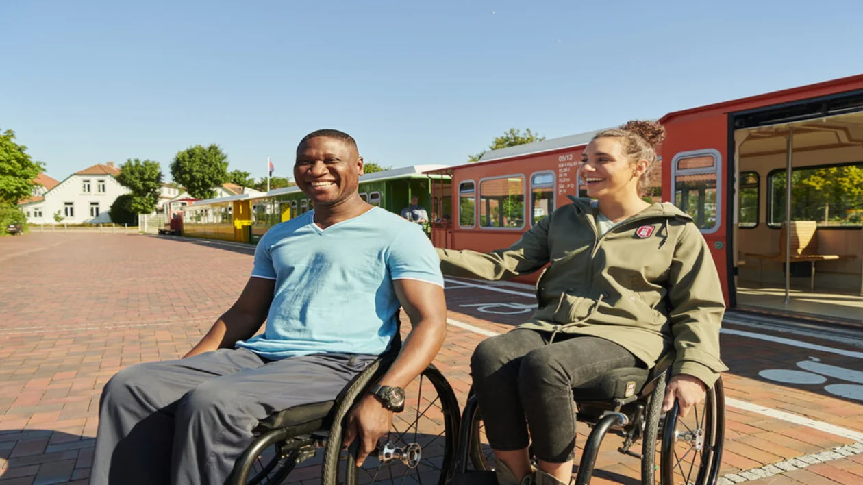 Two people in wheelchairs amiling at a train station on a sunny day.