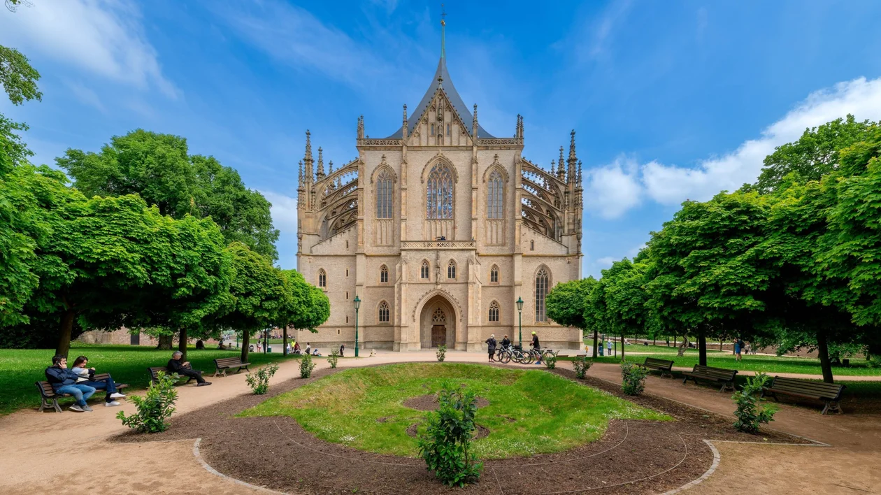 Gothic cathedral with intricate facade, surrounded by green trees and benches under a blue sky.