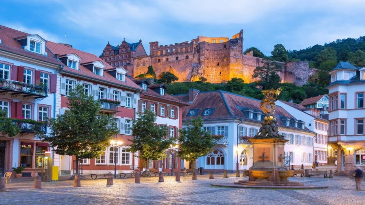 Evening view of Heidelberg's Marktplatz with a glimpse of the Heidelberg Castle illuminated in the background. The cobblestone square, with its charming old buildings and Baroque-style architecture, creates a picturesque atmosphere under the twilight sky.