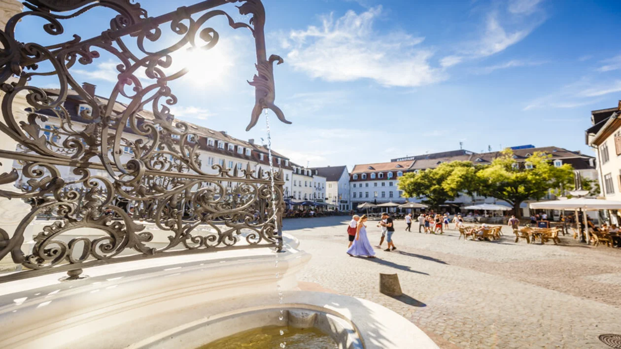 A sunny day in Saarbrücken, showcasing the St. Johanner Markt square. The ornate fountain in the foreground and lively outdoor cafes invite people to relax and enjoy the warmth of the day. Couples and families add to the vibrant, welcoming atmosphere.
