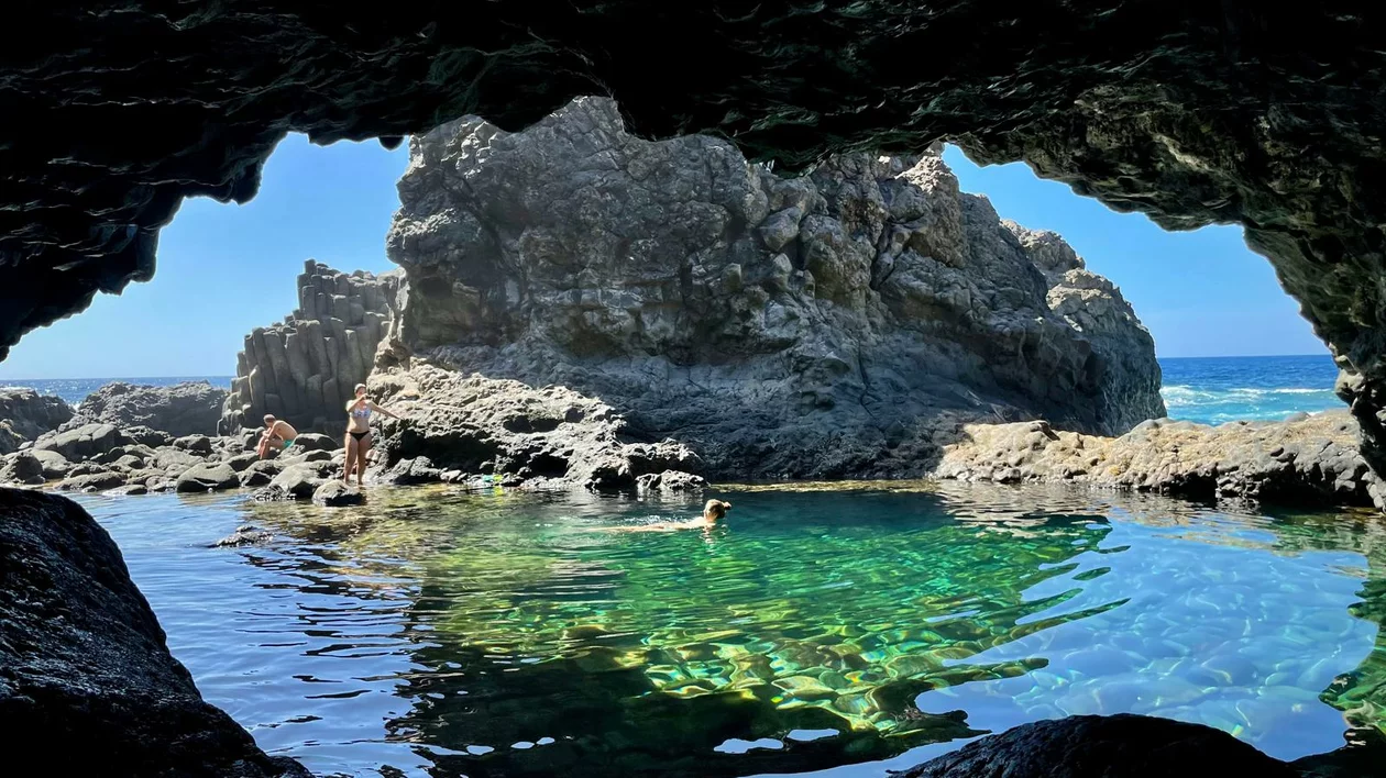 View from a cave of a clear tidal pool surrounded by rocks, with a bright blue sky above.