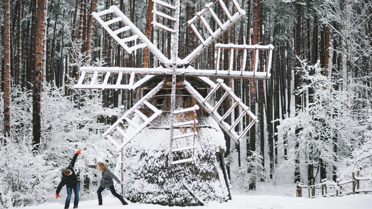 Two kids playing with the snow near a snow-covered windmill in a forest.
