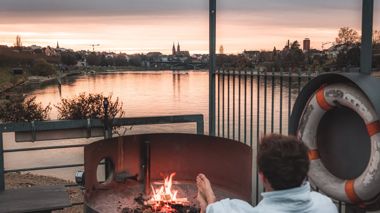 A man on the back sitting outside of a terrace, standing close to a fireplace. On the background, a river and Basel.