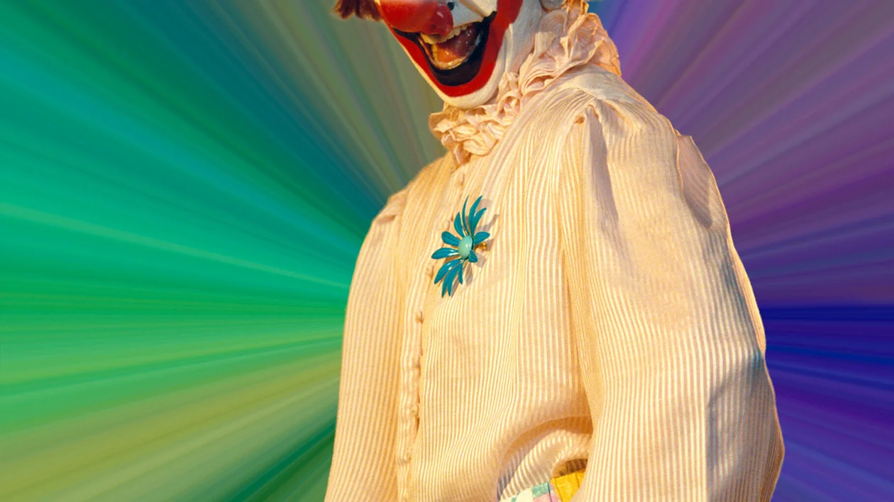 A clown in a striped shirt with a hat and brooch, against a colorful streaked background.