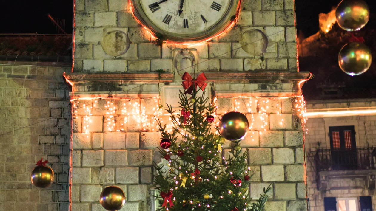 A clock tower decorated with lights. In front of it, a Christmas tree.