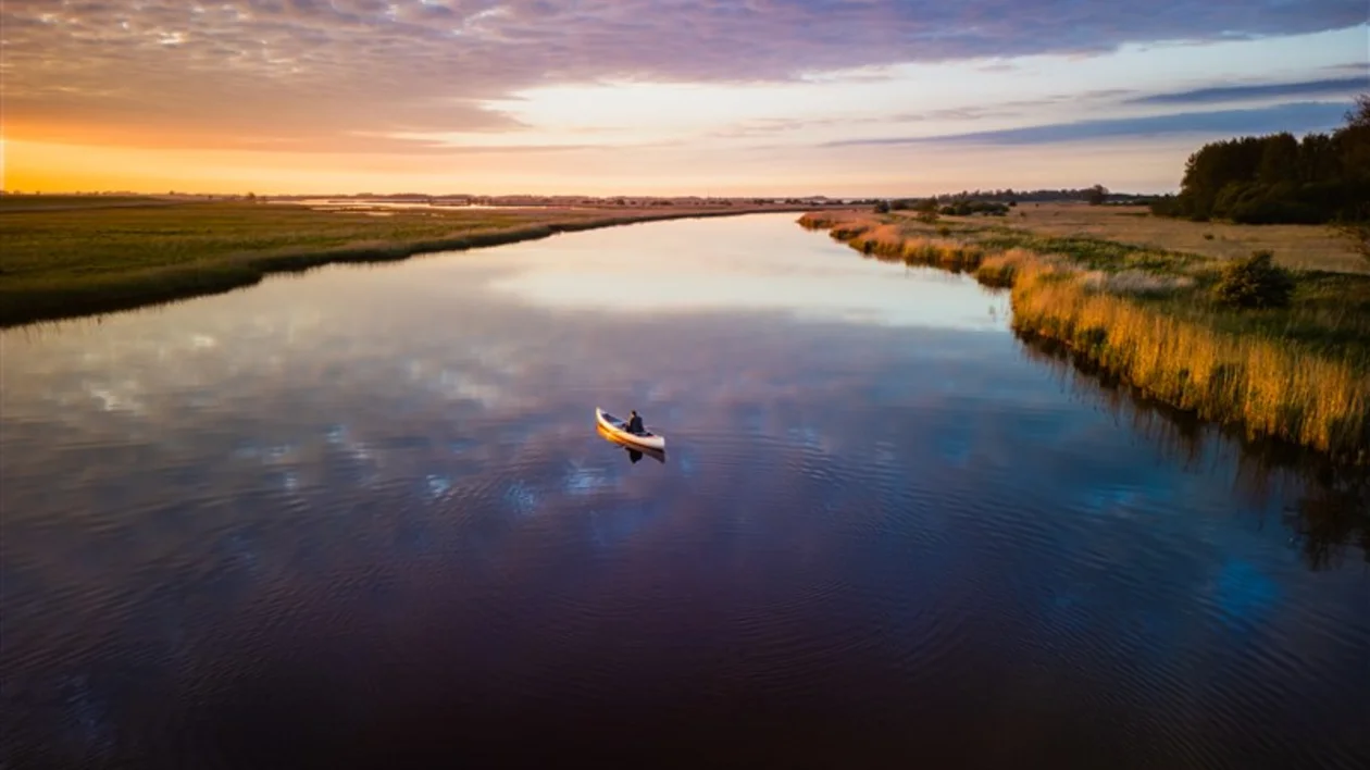 Aerial view of a canoe on a calm river at sunset with clouds reflecting on water.
