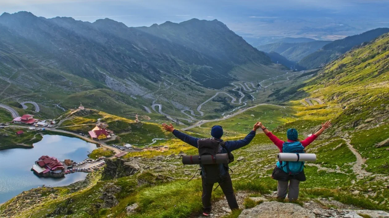 Two hikers with raised arms overlooking a mountain landscape with a winding road and lake.