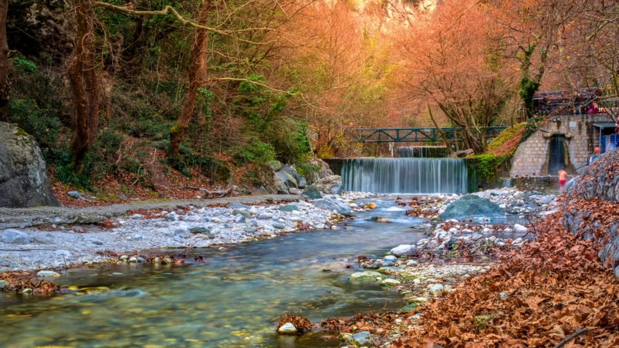 Autumn scenery with a small waterfall, a stream, colorful leaves, and a footbridge.