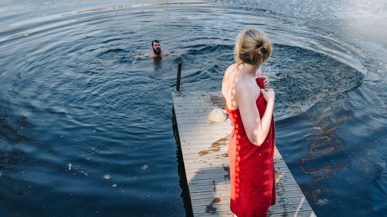 Woman in red towel standing on a wooden dock by the water with a person swimming.