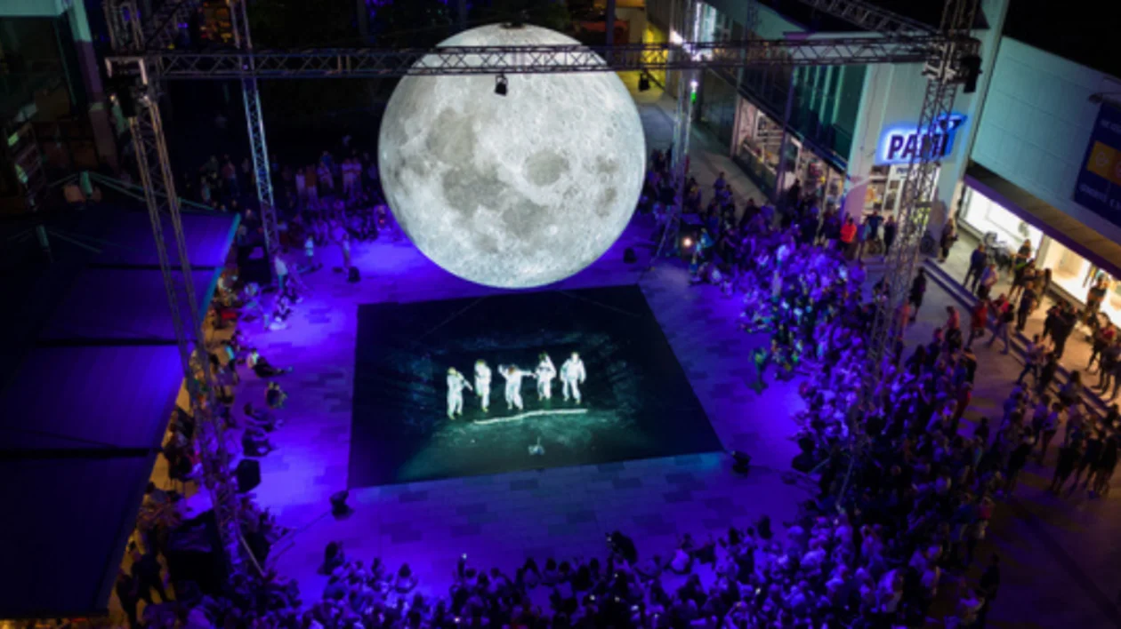 Outdoor event with a large moon replica above performers on stage, surrounded by an audience at night.