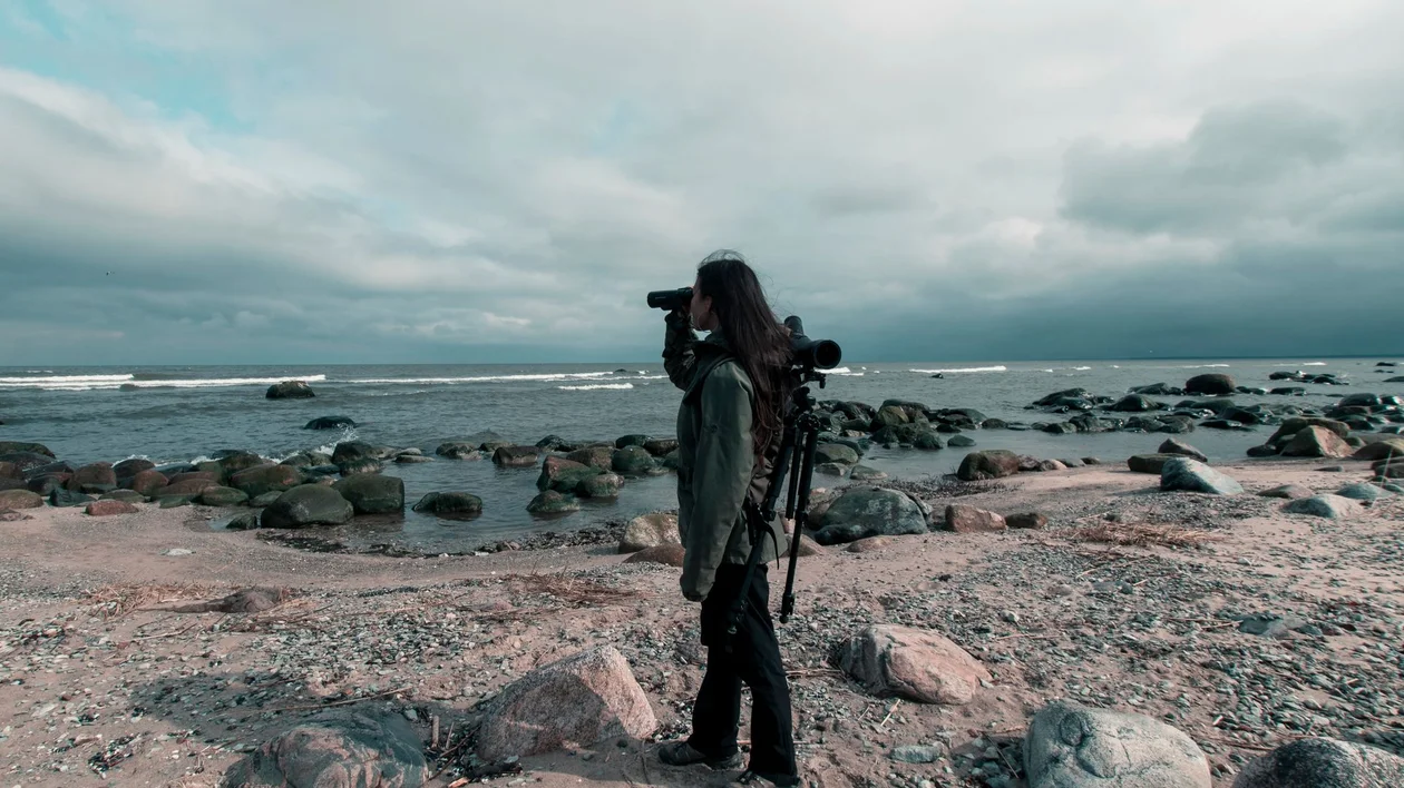 Person with binoculars beside a camera tripod on a rocky shoreline under cloudy skies.