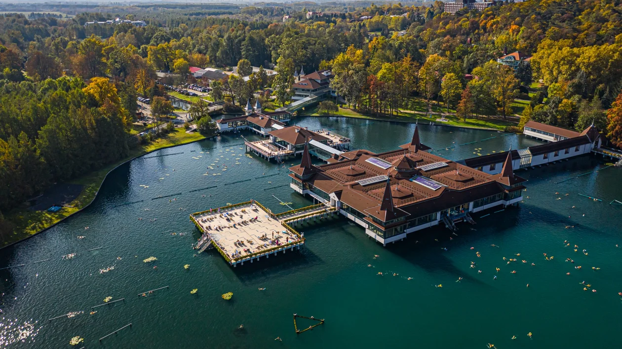 Aerial view of the Hévíz Thermal Lake and its surrounding spa buildings, set amidst lush green foliage and autumn-coloured trees.
