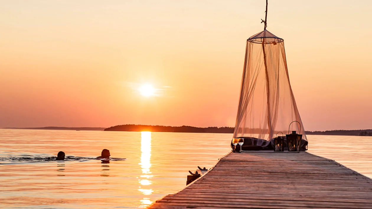 Sunset view from a pier with a mosquito net over a seating area and two people swimming in the lake.