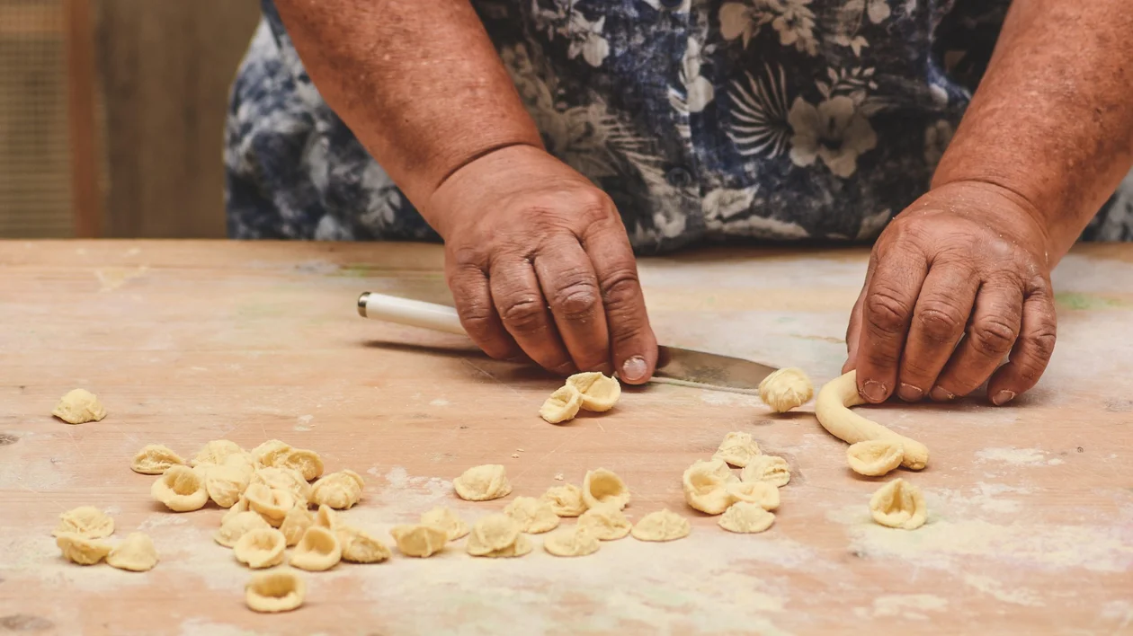 Hands cutting and shaping homemade pasta on a floured wooden surface.