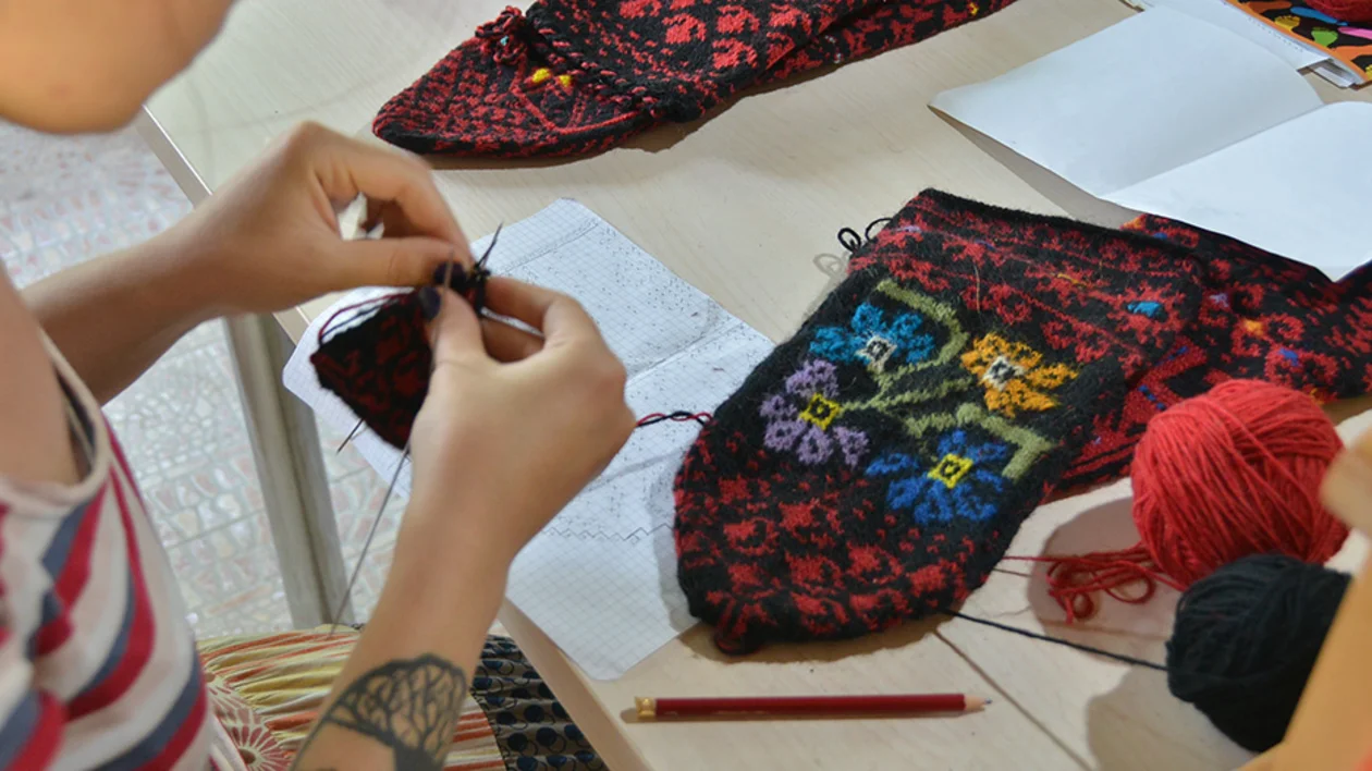 A person crafts colorful socks, surrounded by yarn and sketches, focusing on intricate needlework.