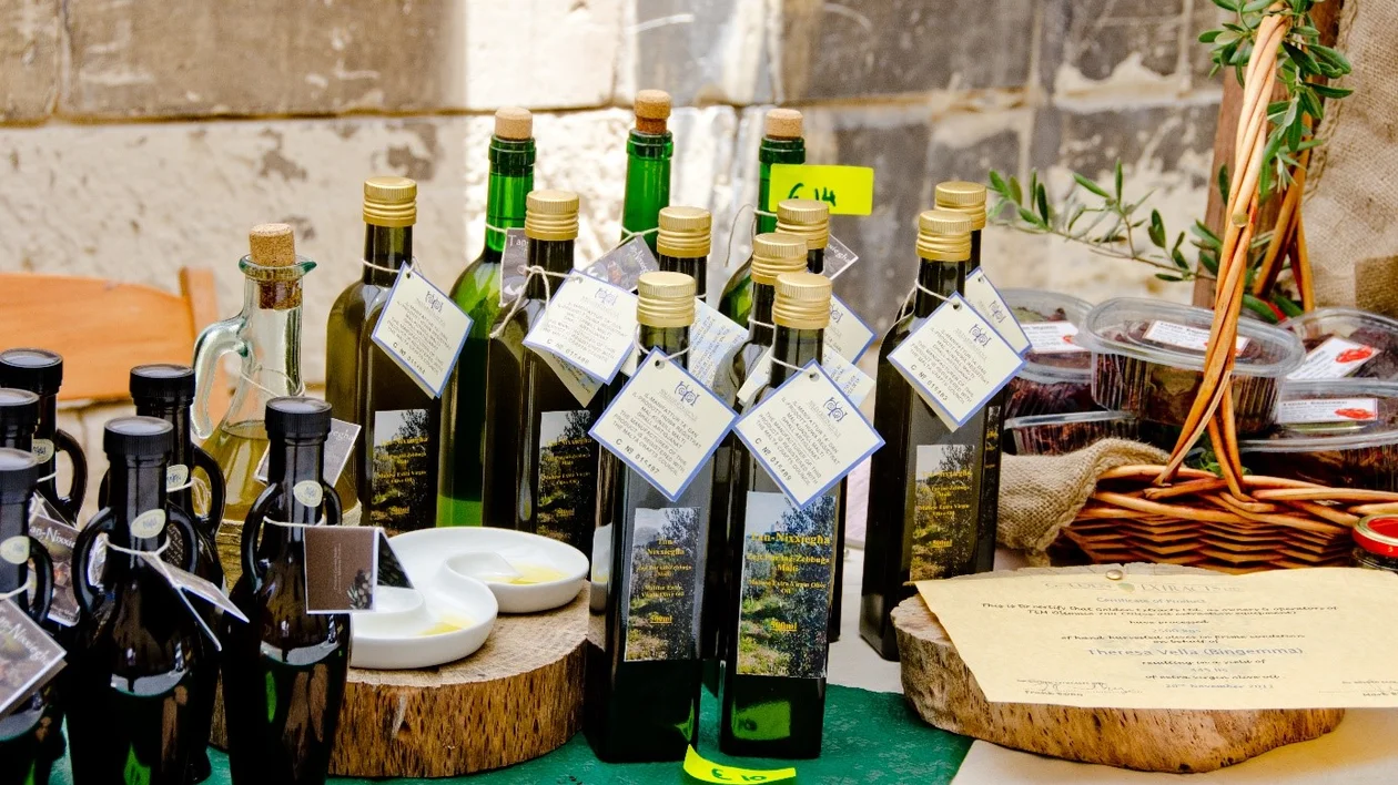 A display of various olive oil bottles with tags, alongside small dishware and a basket of preserves on a green tablecloth.