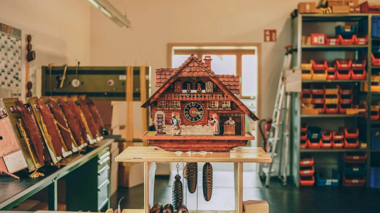A detailed Black Forest-style cuckoo clock with wooden figurines and pinecone-shaped weights, displayed in a traditional workshop.
