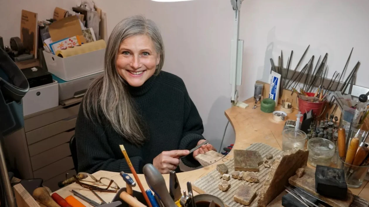 A woman sitting in at a cluttered workbench, surrounded by tools, materials, and projects, focused on their crafting work.