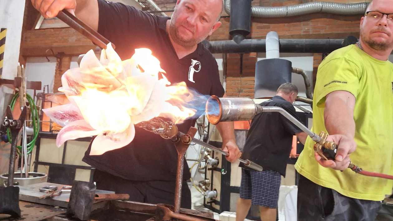 Two artisans work with glass, manipulating molten material with a flame torch in a vibrant glassblowing studio.