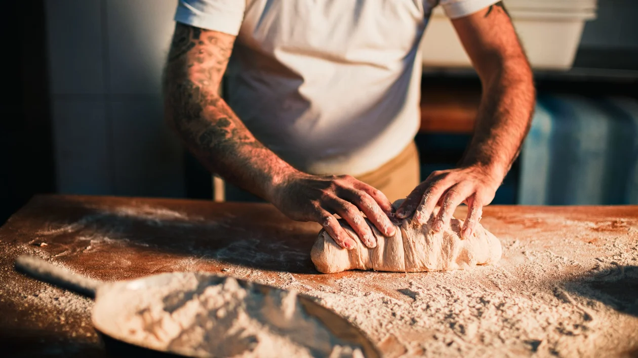 A baker kneads dough on a flour-dusted wooden work surface.