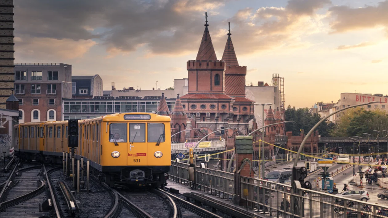 A yellow train navigates tracks near the iconic red-brick Oberbaum Bridge in Berlin during sunset, amidst a bustling urban backdrop.