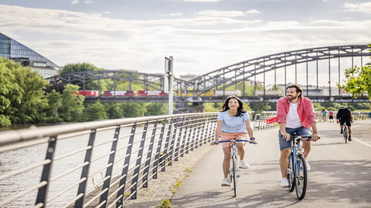 Two cyclists ride along a sunny riverside pathway, with a train crossing a bridge in the background and lush greenery lining the route.