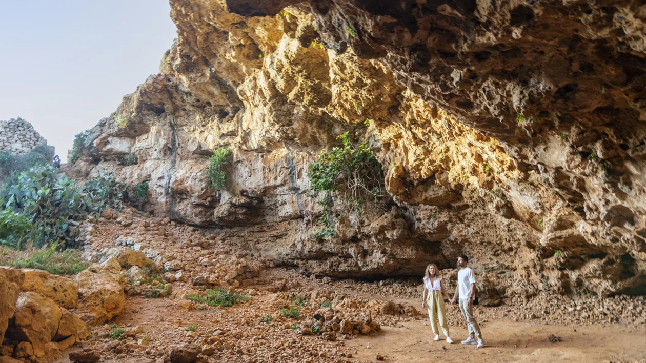 A couple explores a rocky cave, surrounded by textured stone walls and sparse greenery, under a bright sky.