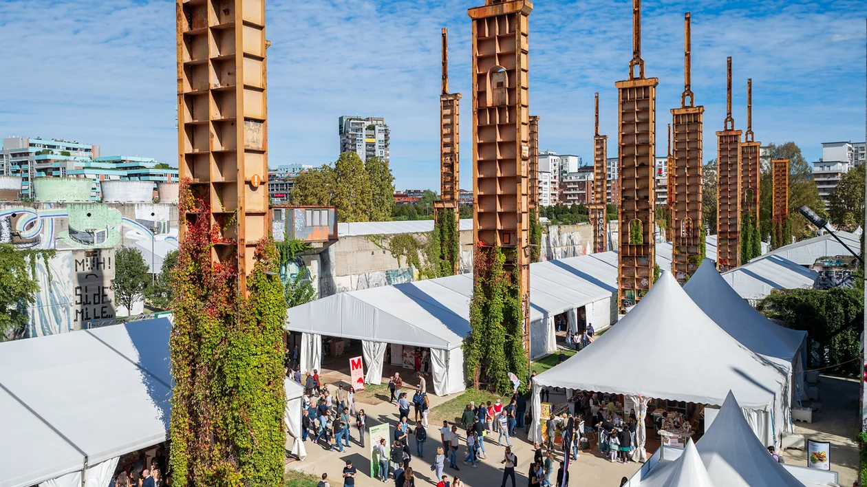 A bustling outdoor event beneath towering rust-colored structures, surrounded by white tents and vibrant graffiti on nearby walls.