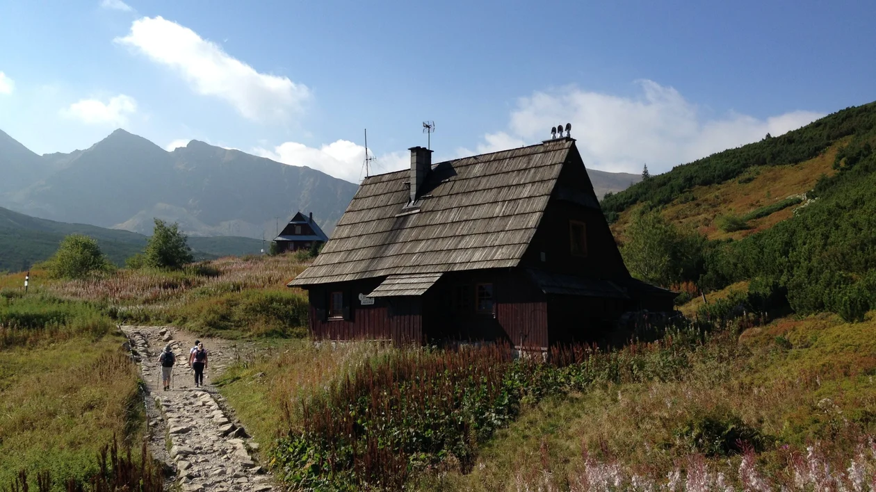 A rustic wooden cabin in a mountainous landscape, with two hikers walking along a rocky path surrounded by wild grasses and hills.