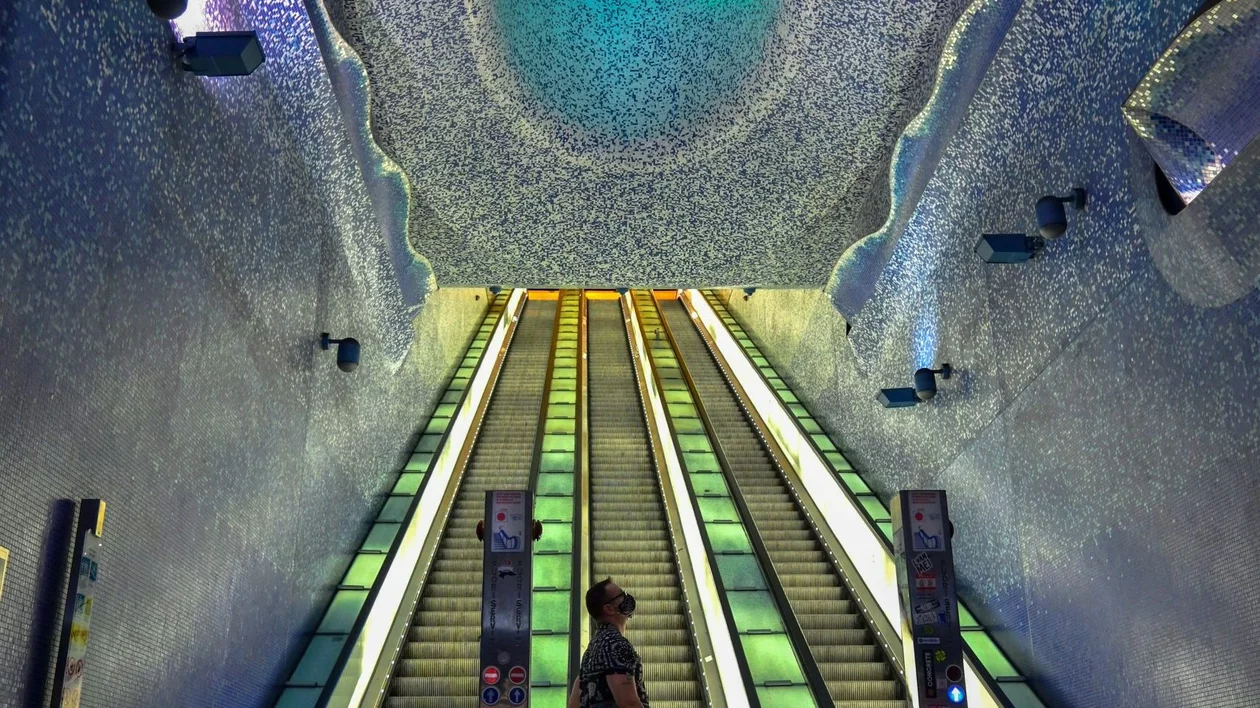 Modern underground station with illuminated escalators and a striking blue mosaic ceiling featuring a large circular light installation.