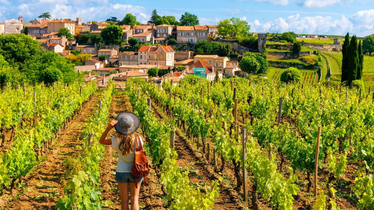 A woman wearing a striped sunhat and backpack walks through a lush vineyard toward a picturesque hilltop village under a bright blue sky with scattered clouds.