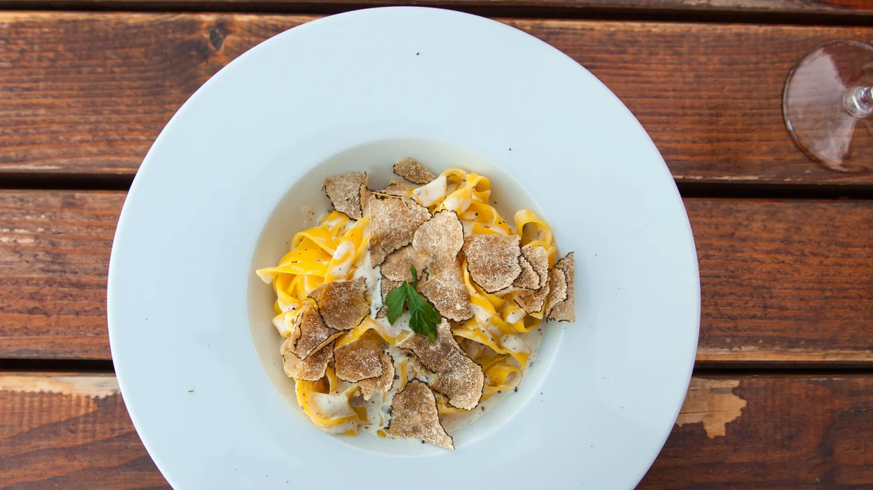A plate of fresh tagliatelle pasta topped with thinly sliced truffles, served on a wooden table, accompanied by a glass of water. The dish highlights the rich and earthy flavors of truffles.