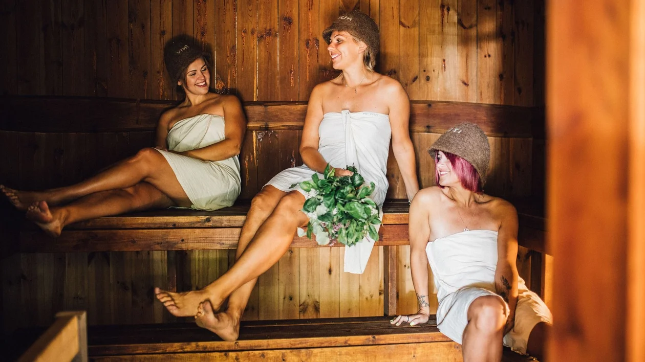 Three women in towels relax in a wooden sauna, with one holding a bundle of green leaves surrounded by a warm, rustic atmosphere.