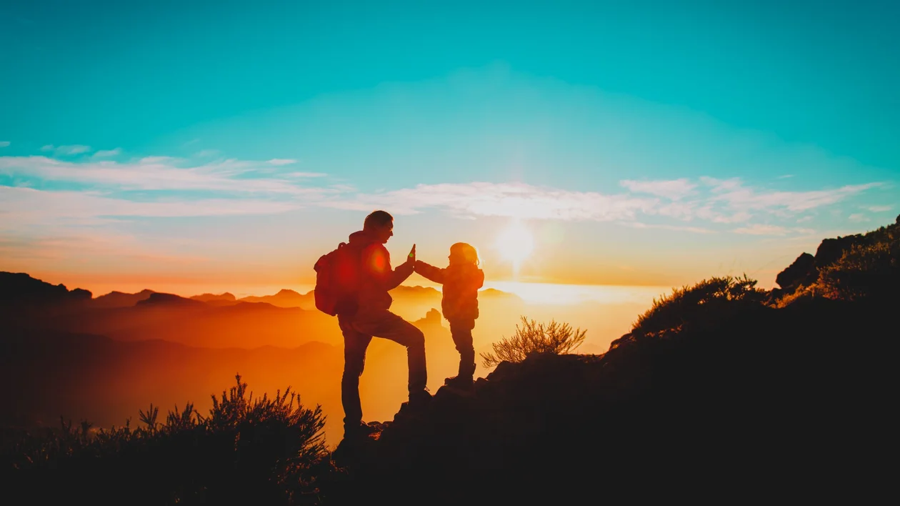 A silhouetted adult and child high-five on a mountain summit at sunset, with dramatic orange light casting over distant peaks and a vibrant blue sky above.