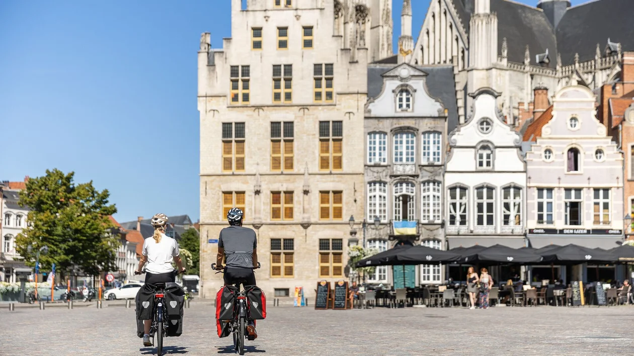 Two cyclists view historic buildings in a vibrant square on a sunny day, with cafés and greenery in the background.