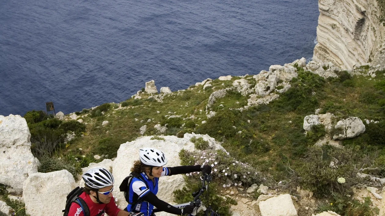 Two mountain bikers push their bikes uphill along a rocky coastal path, with steep cliffs and deep blue sea in the background.
