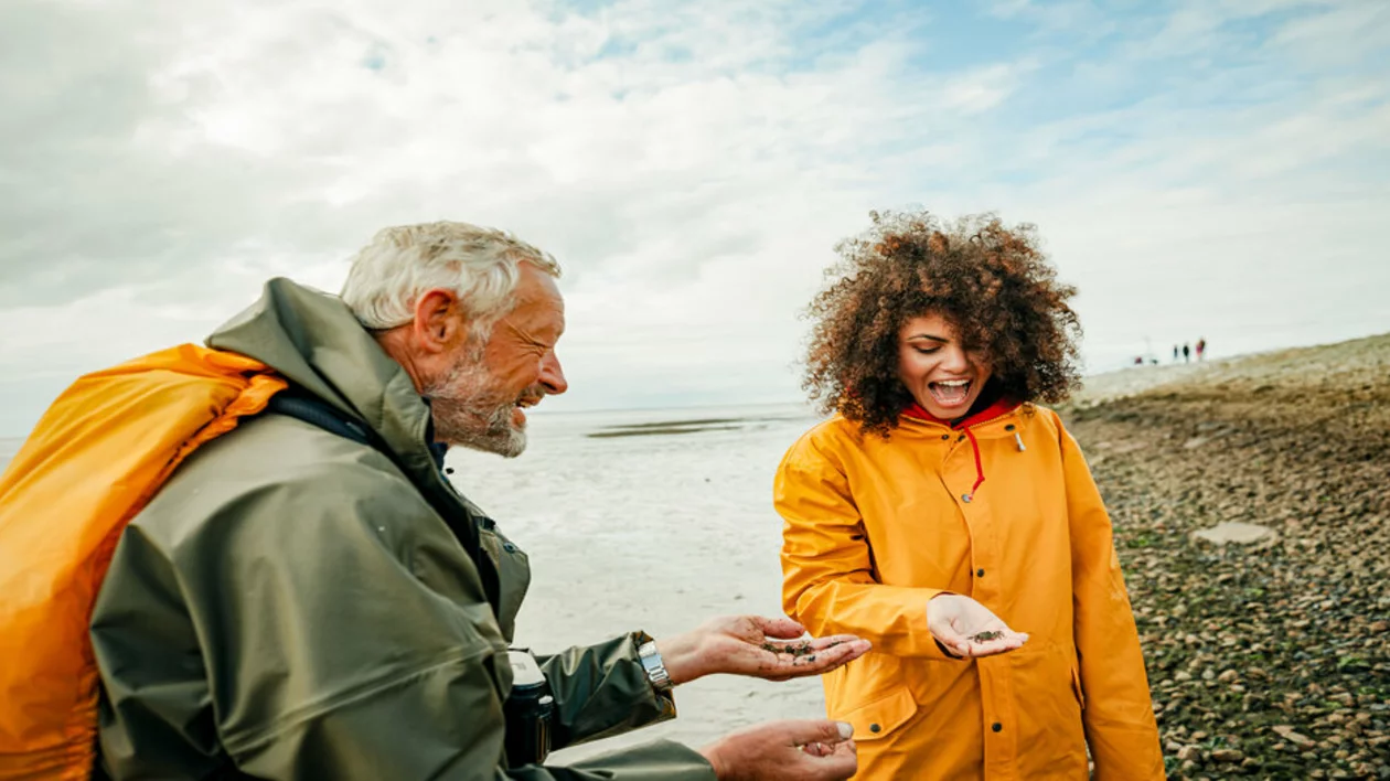 Two people on a rocky beach examine small shells and stones, with cloudy skies and a distant shoreline in the background.
