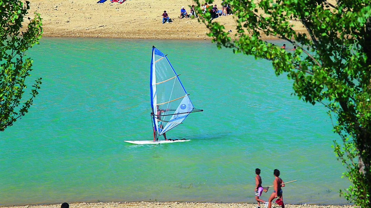 A windsurfer glides across a turquoise lake while sunbathers relax on a sandy shore, kids play nearby, surrounded by lush greenery.