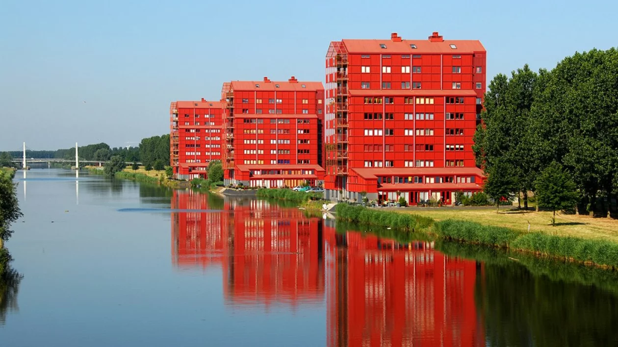 Three red modern apartment buildings reflect in a serene river, surrounded by greenery and a clear blue sky.