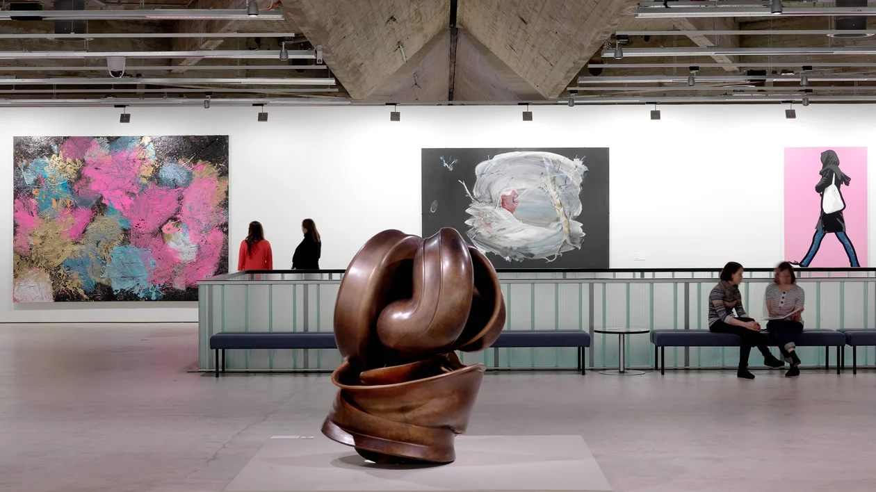 A contemporary art gallery showcasing a bronze sculpture in the foreground and colorful abstract paintings on the walls.
