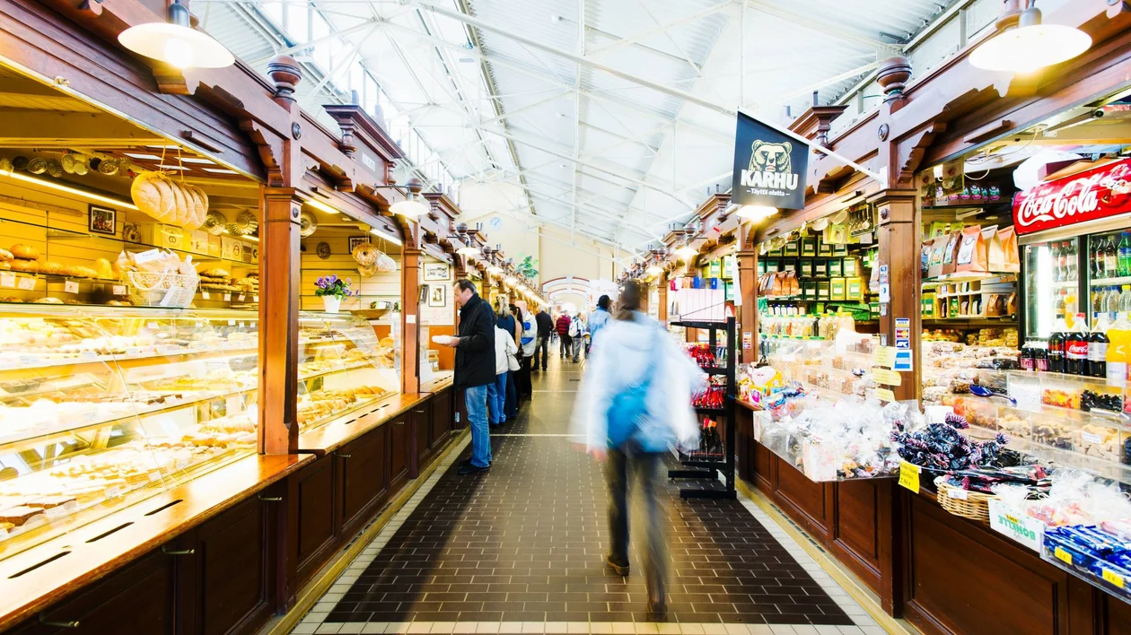 A bustling market interior with wooden stalls filled with baked goods and snacks, and shoppers exploring the vibrant offerings.