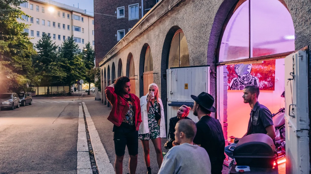 A group of stylish individuals converse outside a venue with colorful lighting, set against an urban backdrop.
