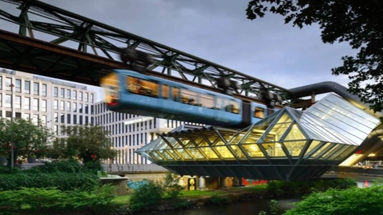 A suspended blue train glides above a modern glass building, set against a backdrop of urban architecture and greenery.