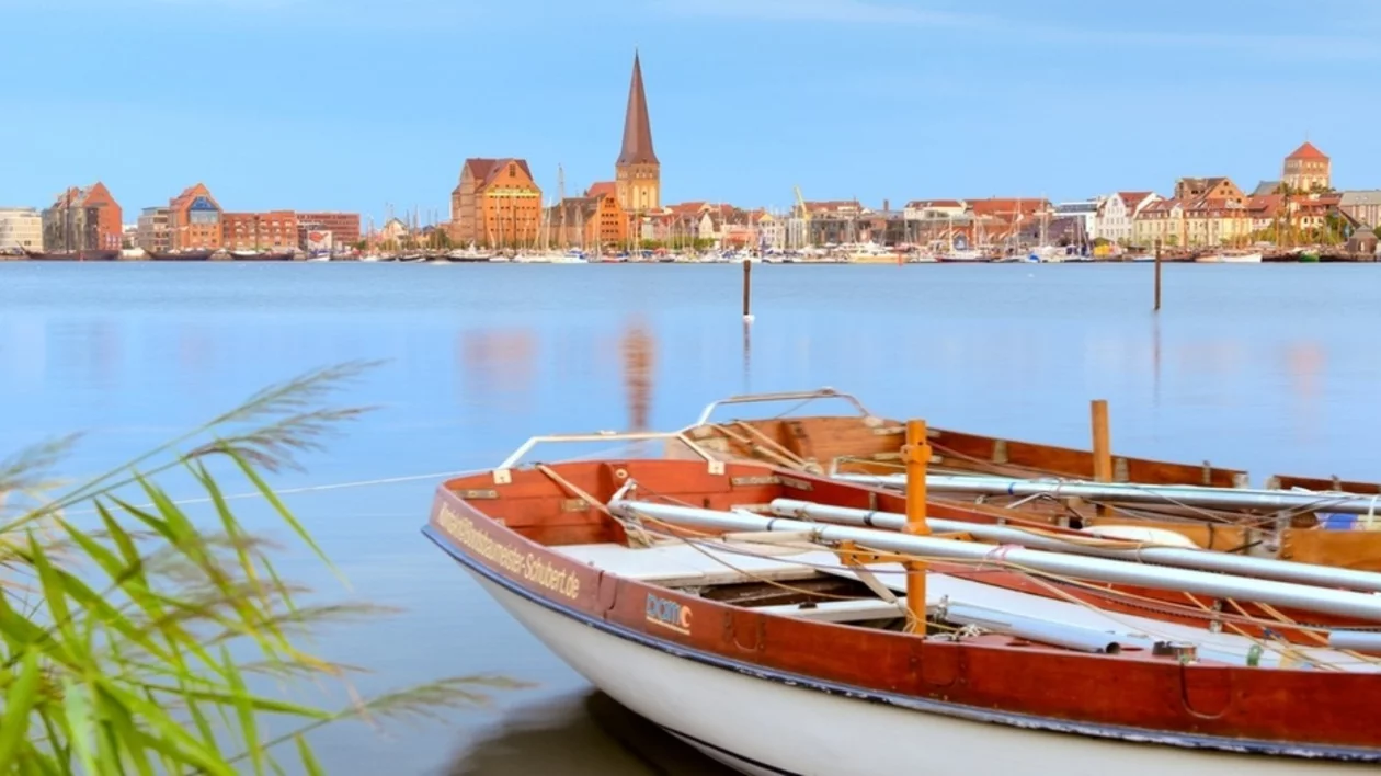 Serene lakeside view with rustic boats in foreground, featuring a skyline of colorful buildings and a church steeple in the background.