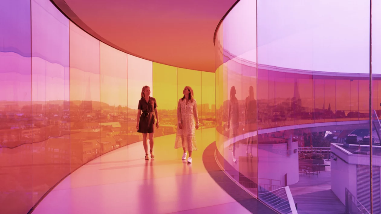 Two women stroll through a brightly colored glass walkway, reflecting a vibrant urban landscape in shades of purple, orange, and yellow.