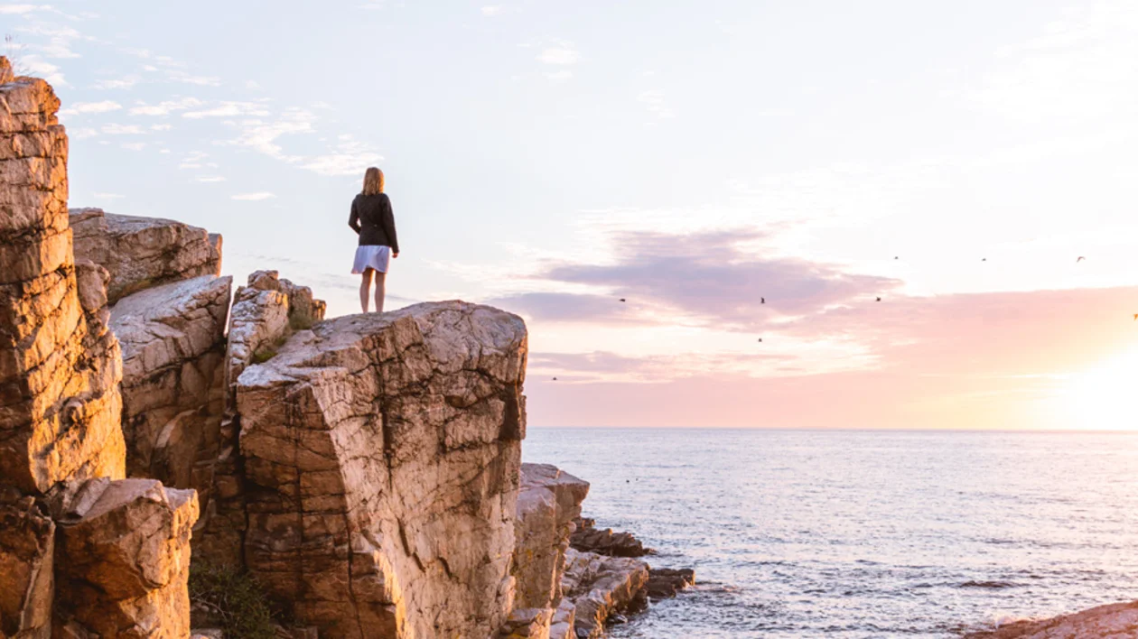Girl standing on coastal rocks at Gudhjem, Bornholm, Denmark, admiring the sea at sunset.