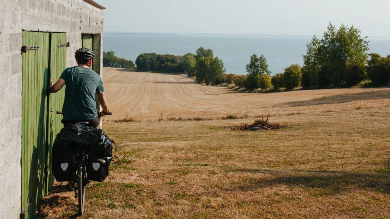 Cyclist departing from a green-doored shed, overlooking fields and the sea in Bølgemose, Ærøskøbing, Denmark.