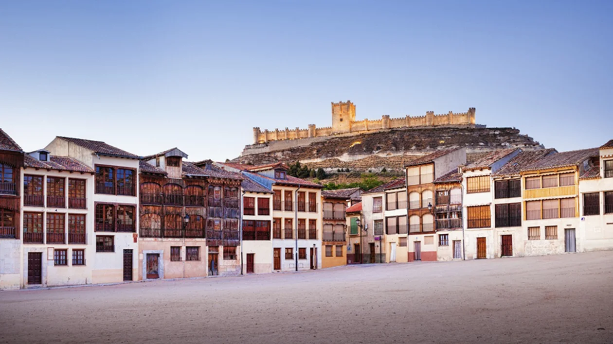 Historic wooden buildings line a plaza, with a castle perched atop a hill in the background under a clear blue sky.